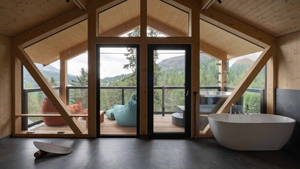 A modern interior with a large bathtub, a wooden deck with seating, and a view of a mountainous landscape. - Deluxe chalet with sauna & hot tub (Turrach)