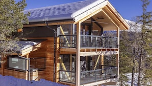 Una cabaña de madera con balcón y ventanas amplias, rodeada de nieve y árboles.