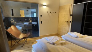 Ein modernes Hotelzimmer mit zwei Betten, Holzboden, Glastrennwand, einem Sessel und einem durch die Glaswand einsehbaren Badezimmer.