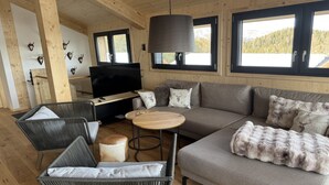 Ein gemütliches Blockhaus-Interieur mit einer Couch, Sesseln, einem Fernseher und einem großen Fenster mit Blick auf die Berge.