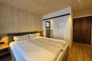 A bedroom with a wooden ceiling, a bed with white bedding, a bedside table, a mirror, and a wooden door. - Superior chalet in Turrach with sauna (Turrach)