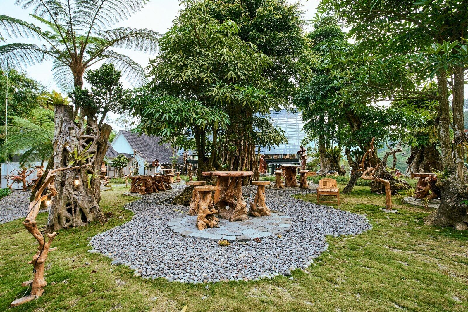 Um jardim com mesas e cadeiras de madeira, cercado por árvores e um edifício ao fundo.