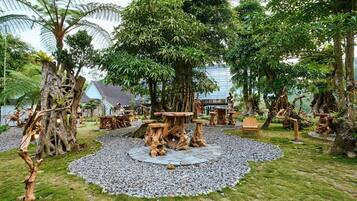 Um jardim com mesas e cadeiras de madeira, cercado por árvores e um edifício ao fundo.