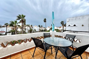 Une terrasse avec une table et des chaises en verre, un parasol bleu et une vue sur des palmiers et des immeubles.