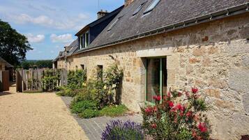 Un bâtiment en pierre avec une allée gravillonnée et un jardin agrémenté de fleurs et d’arbustes.