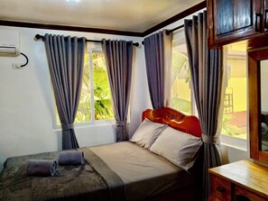 Una camera da letto con un letto, tende, una finestra vista verde, una testiera in legno e un comodino.