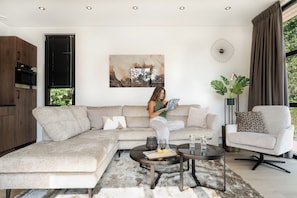 A modern living room with a sectional sofa, a coffee table, a side table, a potted plant, and a large abstract painting on the wall. - Luxurious 4-person wellness villa on the Vogelenzang Estate with a private sauna. Bright, comfortable, and sustainably furnished, surrounded by greenery and close to the dunes and coast. (Vogelenzang)