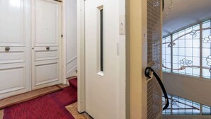 Een gang met dubbele deuren, een rood tapijt, een klein raam met een metalen rooster en een trap met een decoratieve balustrade.