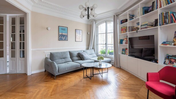 Een woonkamer met een grijze bank, een ronde salontafel, een rode fauteuil, een boekenkast en een televisie.