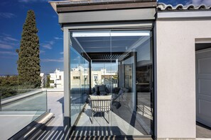 Een modern gebouw met een glazen gevel, een terras op het dak met buitenmeubilair, en een grote boom op de achtergrond.