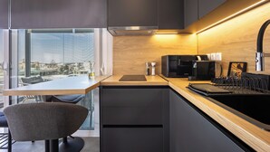Cozinha moderna com fogão embutido, micro-ondas e máquina de café. Há uma mesa de jantar com cadeiras e vista para a cidade através de amplas janelas.