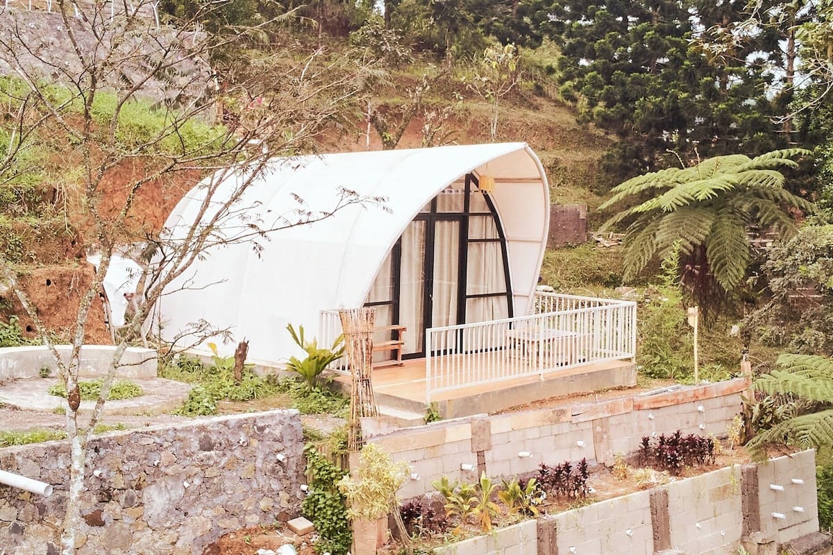 Une structure en forme de dôme, dotée d’un balcon, entourée de végétation et d’un mur de pierre.