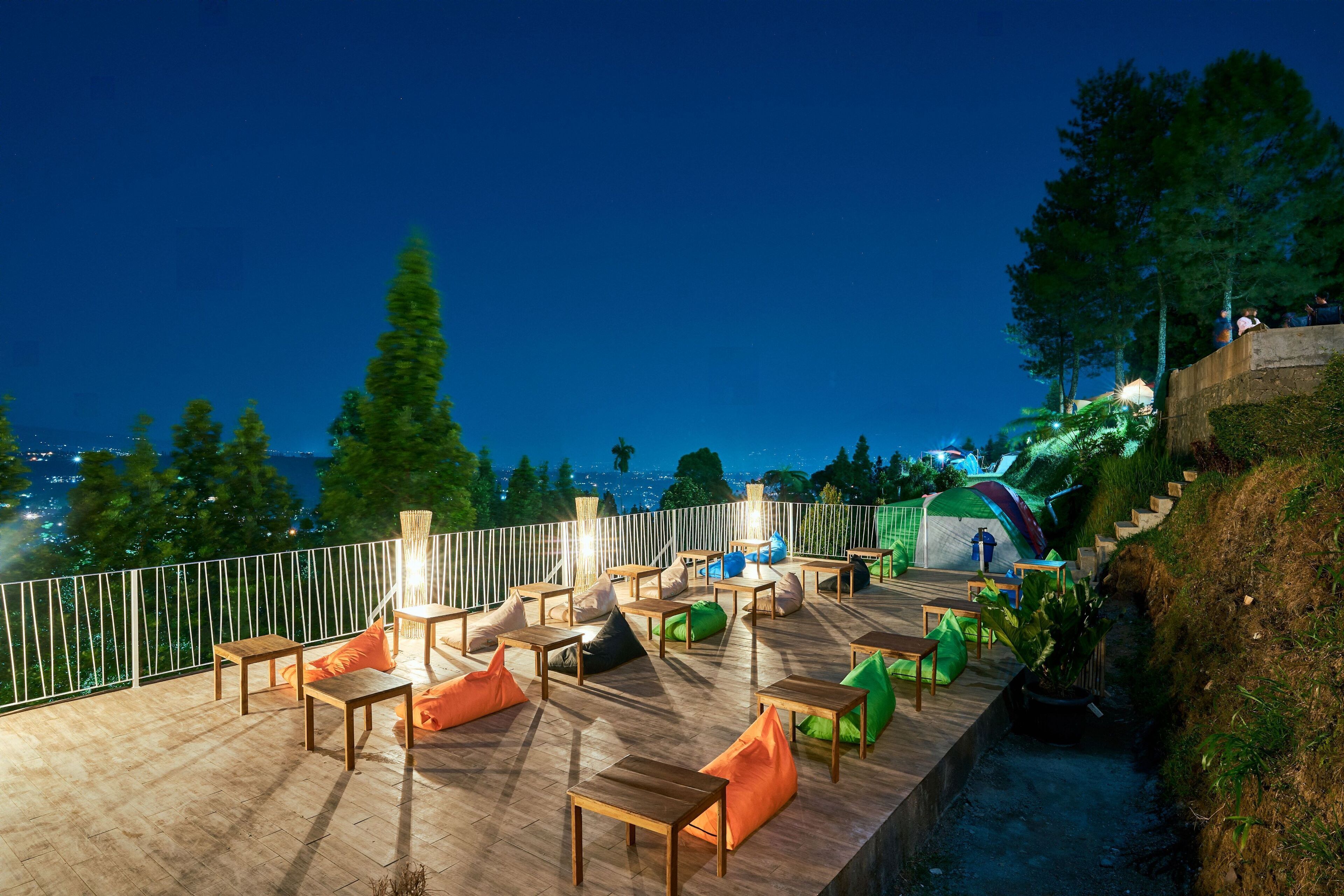 An outdoor seating area with tables and chairs, a grassy slope, and a view of the city at night.