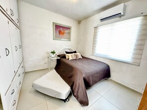 A bedroom with a bed, a bedside table with a plant, a wardrobe, a window with blinds, and an air conditioning unit.