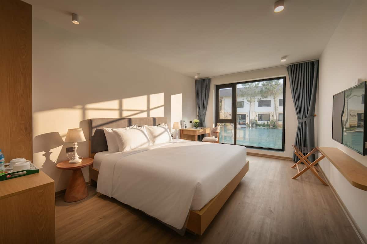 A modern hotel room with a large bed, a desk, a chair, a TV, and a view of a pool.