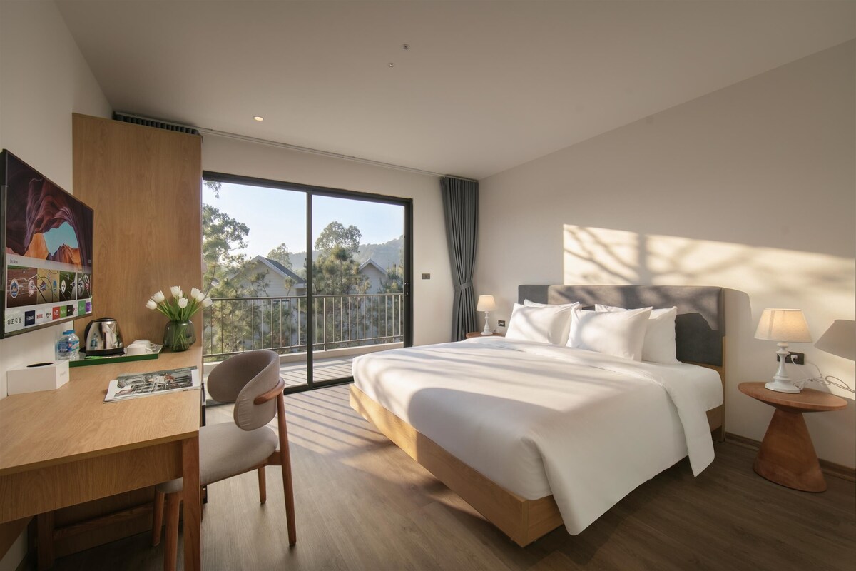 A modern hotel room with a large bed, a desk with a chair, a small table with a lamp, and a view of the outdoors.
