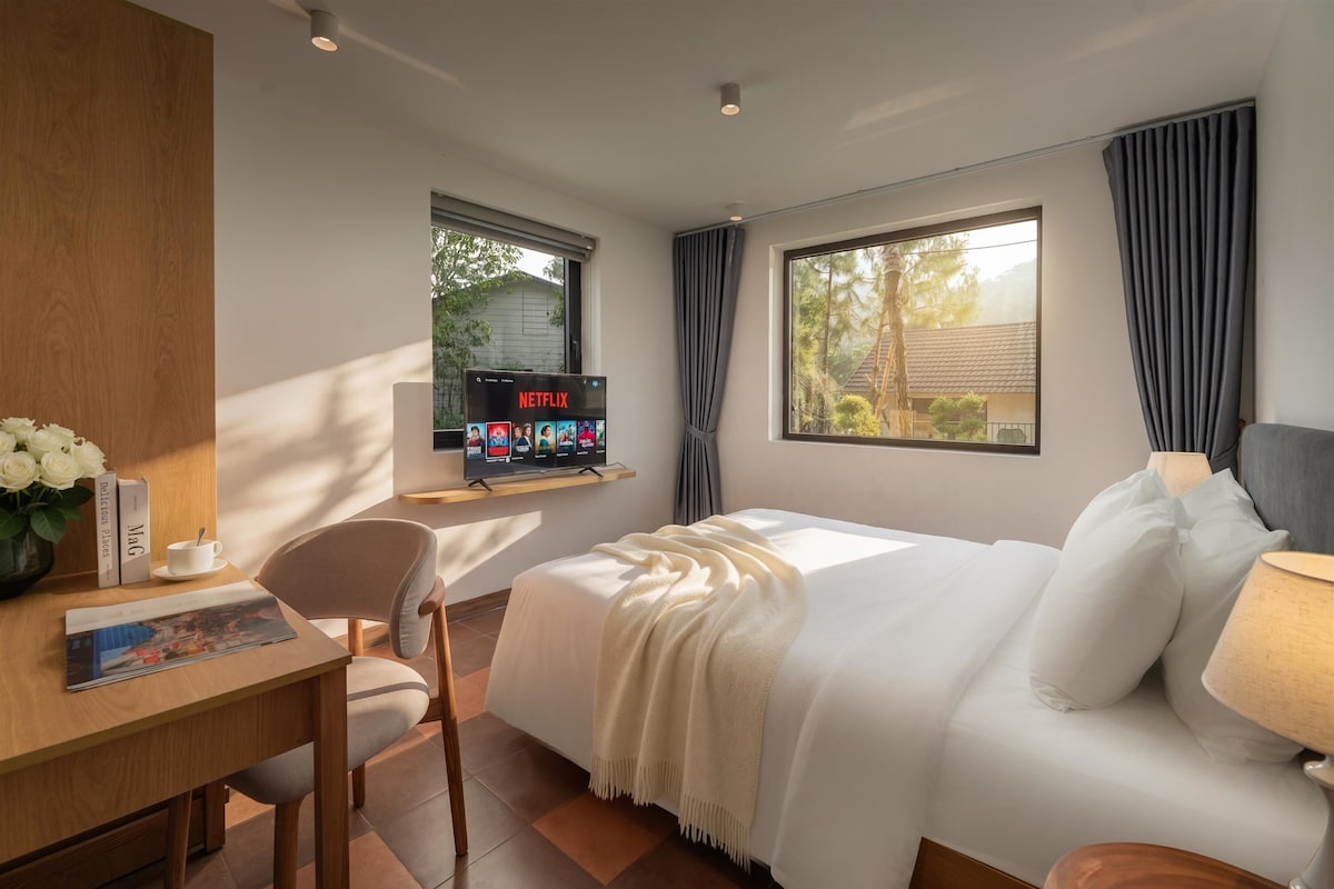 A hotel room with a bed, a desk, a chair, a TV on a stand, and a window with curtains.