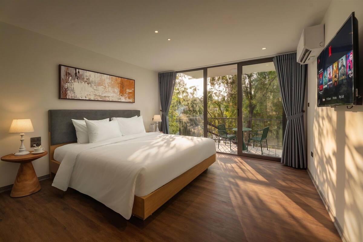 A hotel room with a bed, a TV, a painting, and a balcony with chairs.