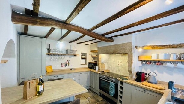 A compact kitchen with exposed wooden beams, a central island, and a built-in oven. - Peak District Retreat · Hope Valley · Dogs Welcome (Stoney Middleton)
