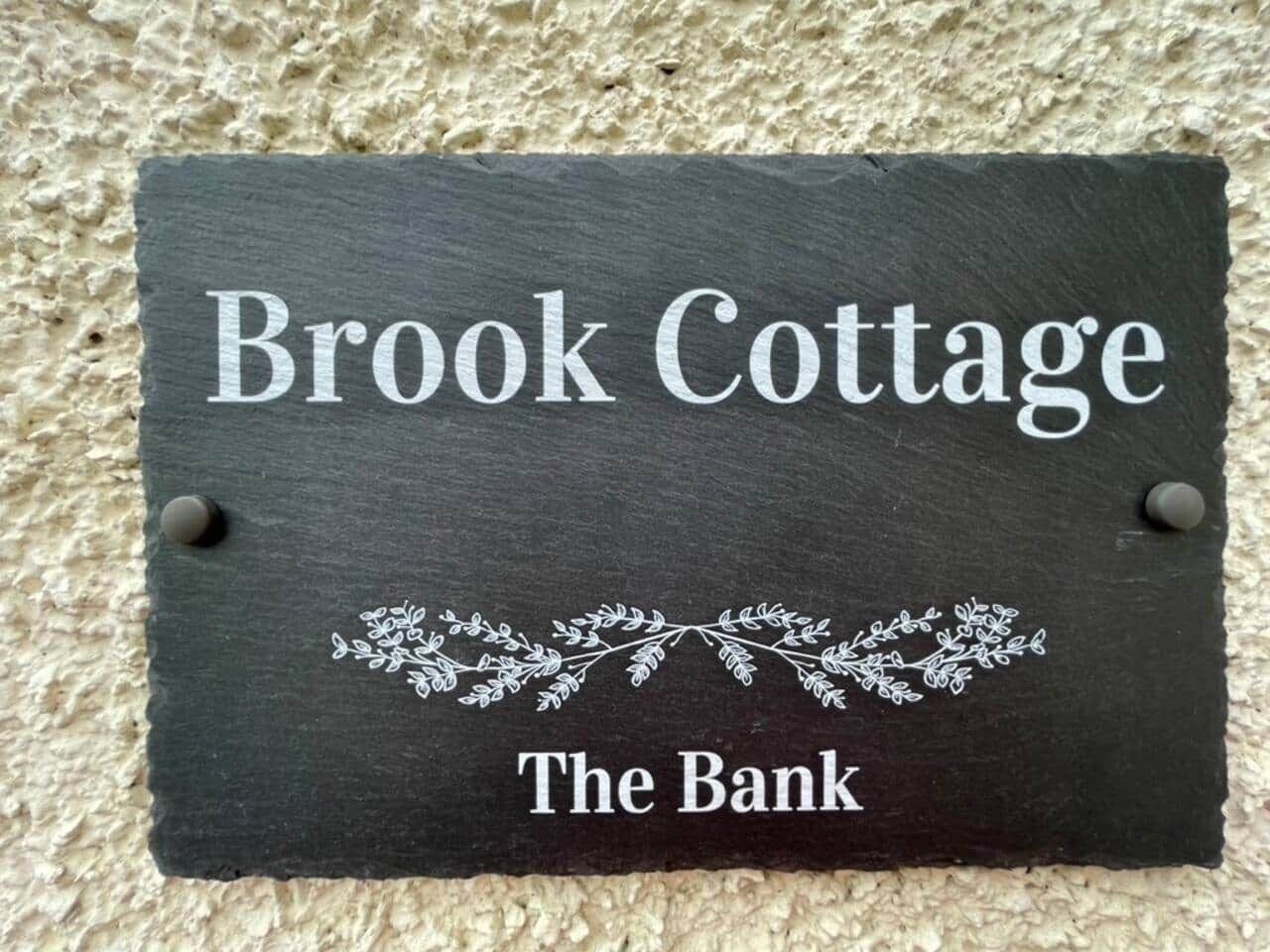 Ein Schiefer Schild mit der Aufschrift 'Brook Cottage The Bank', befestigt an einer strukturierten Wand.
