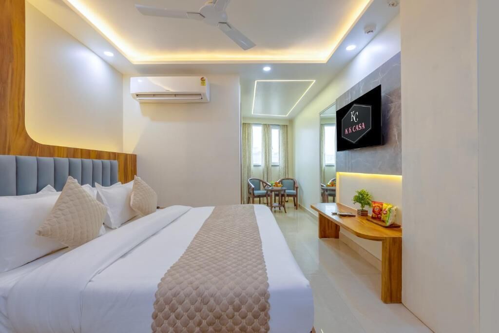 A modern hotel room with a bed, bedside table, TV, and a small seating area with chairs.