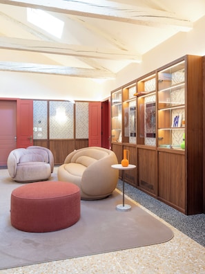 Un interior moderno con una zona de estar que incluye un pouf redondo, un sofá color beige y una silla roja. Un mueble de madera con puertas de cristal exhibe diversos objetos.