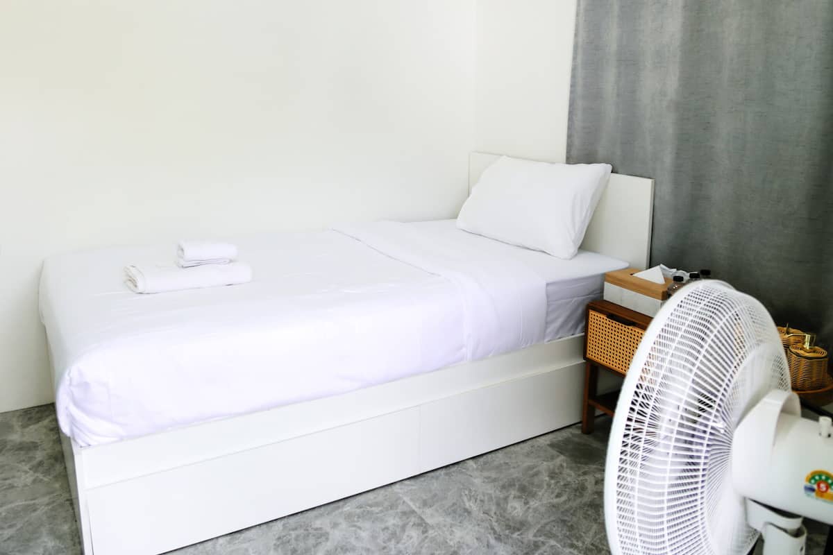 A white bed with a pillow, a white fan, and a small wooden table with a basket.