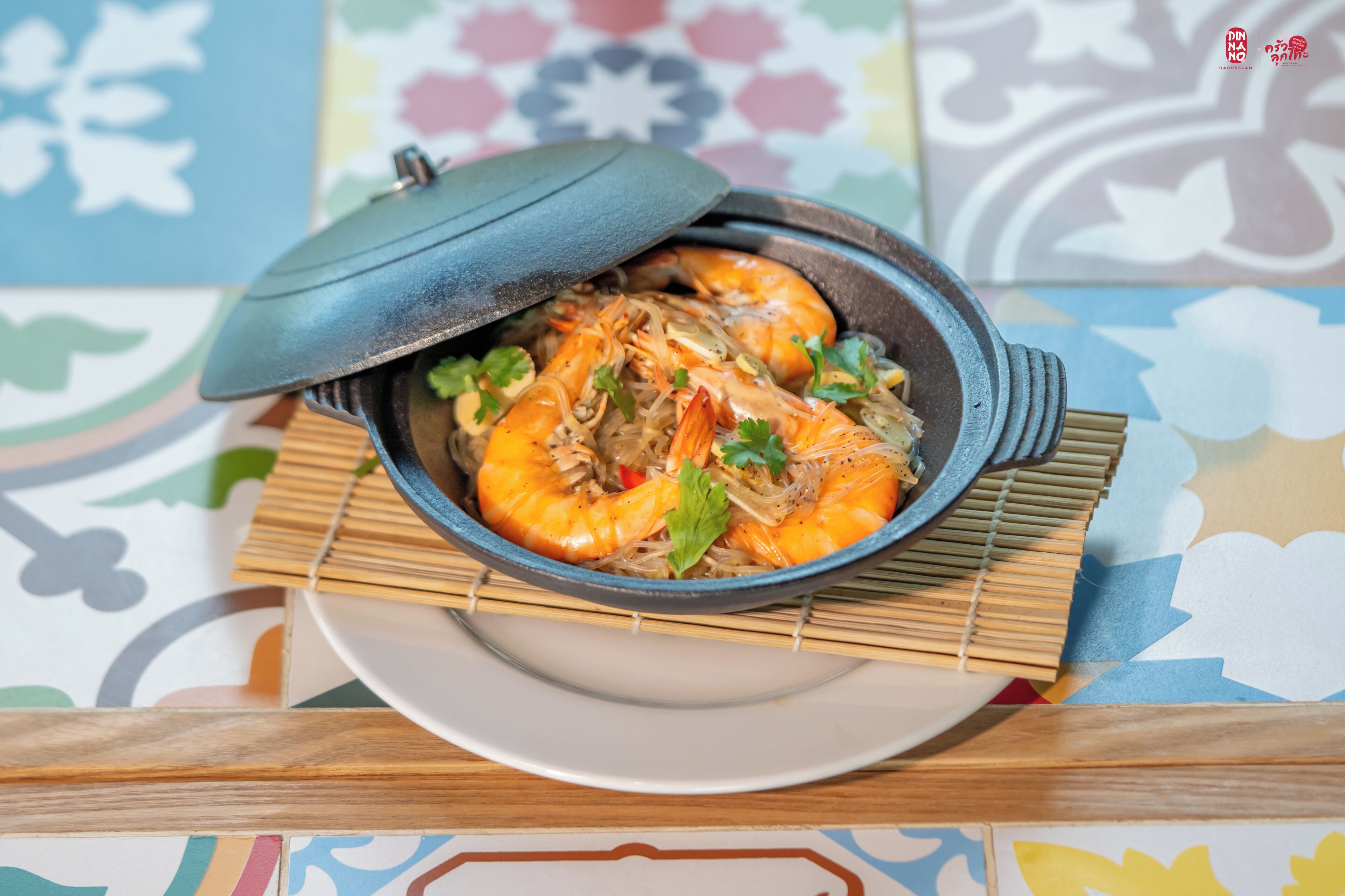 Un bol de camarones cocidos con una tapa, sobre un plato con una estera de bambú debajo.