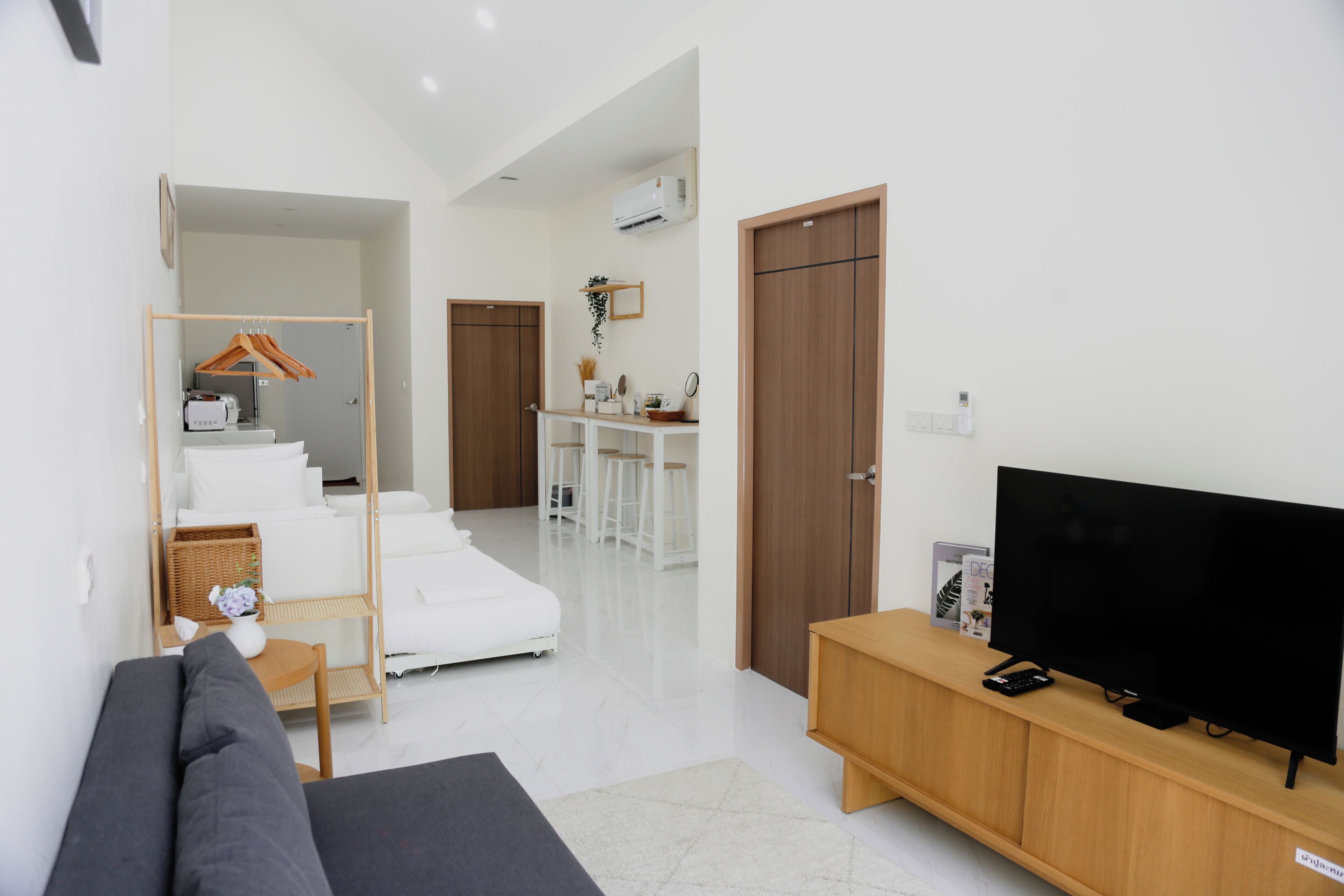A modern living room with a sofa, a TV on a wooden stand, a dining area with stools, and a kitchen area with a hanging plant.