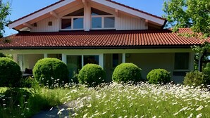 Ett tvåvåningshus med ett rött tegeltak, vita fasadväggar och en täckt veranda. Framför huset finns välskötta buskar och en trädgård med vita blommor.