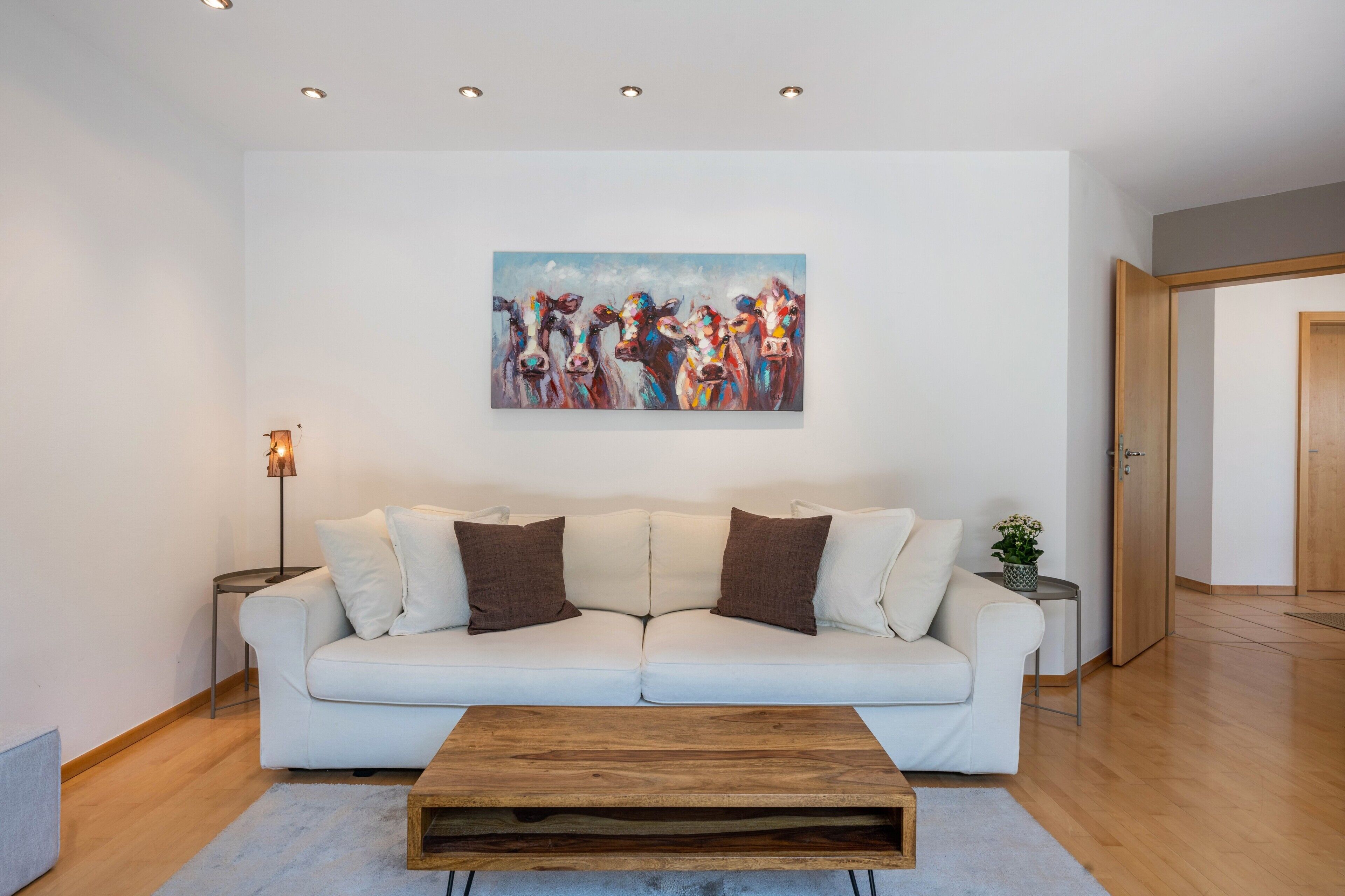Ein modernes Wohnzimmer mit einem weißen Sofa, einem Holztisch und einem Bild von Kühen an der Wand.
