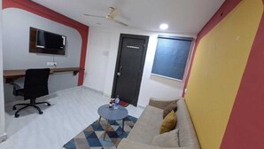 A room with a TV mounted on the wall, a ceiling fan, a door, a sofa, a small table with a bottle, and a chair. - Urban Stay Home (Pune)