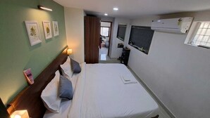 A bedroom with a bed, bedside table, wall-mounted air conditioner, and framed pictures on the wall. - Urban Stay Home (Pune)