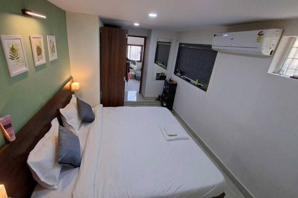 A bedroom with a bed, bedside table, wall-mounted air conditioner, and framed pictures on the wall.