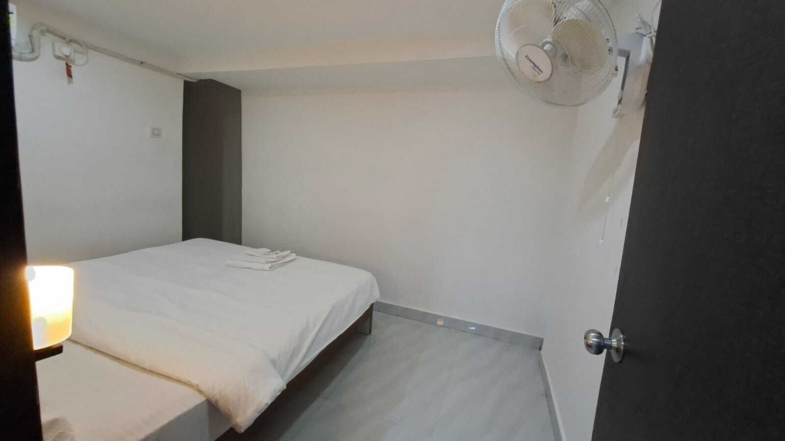 A small, single-bed room with a bedside table, a wall-mounted fan, and a door.
