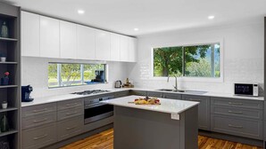 Une cuisine moderne avec des meubles blancs, un îlot central, une cuisinière intégrée et une fenêtre donnant sur un espace verdoyant.