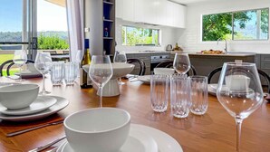 Une cuisine moderne avec des meubles blancs, une table à manger dressée avec de l’assiette blanche, des bols et des verres à vin, et une vue sur la verdure à l’extérieur.