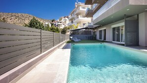 Una piscina amb tanca de fusta, un edifici amb balcons i un turó al fons.