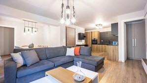 Un saló modern amb un sofà seccional gris, una taula baixa de fusta i una cuina amb armaris de fusta i taulell negre.