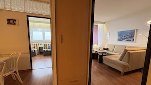 Ett rum med trägolv, en vit stol, en soffa, ett bord med en lampa och ett fönster med gardiner.