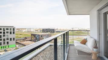 Ein Balkon mit einer weißen Couch und einem Blumentopf, mit Blick auf eine Stadtlandschaft aus modernen Gebäuden und einem klaren Himmel.