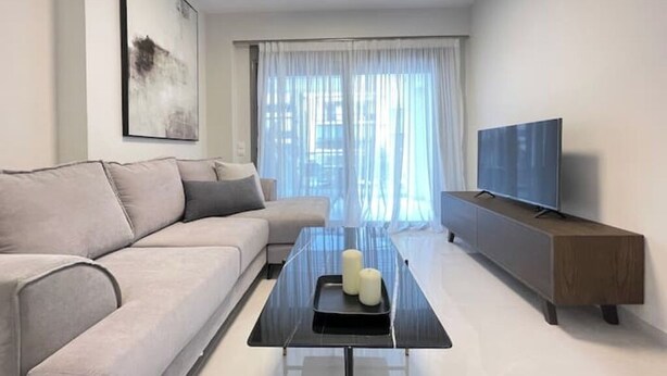 Ein modernes Wohnzimmer mit einer Couch, einem Couchtisch mit Kerzen, einem Fernseher auf einer Holzkommode und einem großen Fenster mit transparenten Vorhängen.