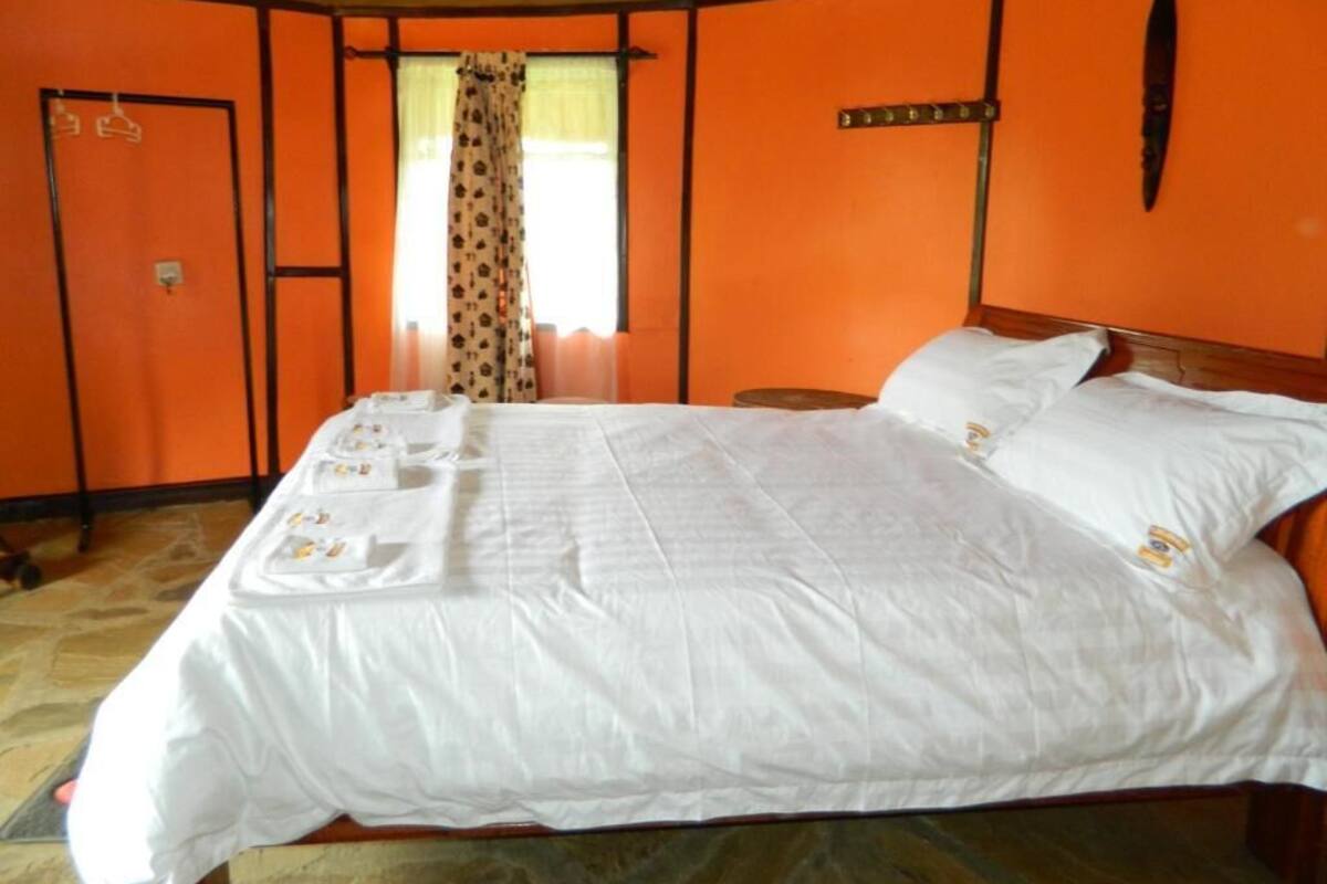 Una habitación con una pared naranja, una cama con ropa blanca, un cabecero de madera y una mesita redonda pequeña.