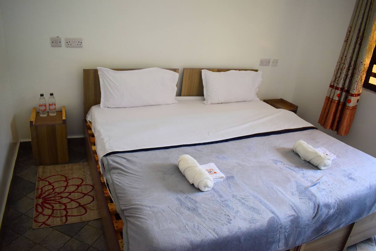 Habitación de hotel con una cama, dos almohadas, una mesita de noche con botellas y una ventana con cortinas.