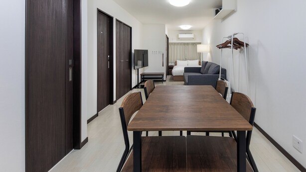 Una habitación de hotel moderna con una mesa de comedor de madera, un armario de madera oscura, un sofá gris y un televisor.