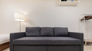 Un sofá gris con tres cojines, una lámpara de pie y un perchero en una habitación con paredes blancas.