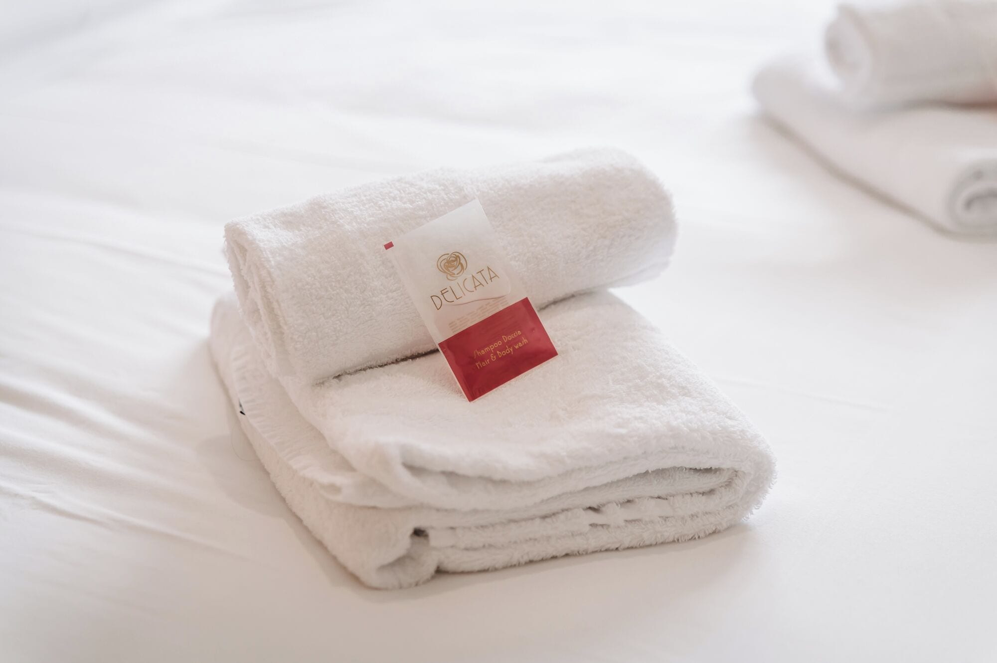 A neatly folded white towel with a tag labeled 'DELICATA' and a smaller red tag indicating 'Shampoo Dolce hair & body wash.'