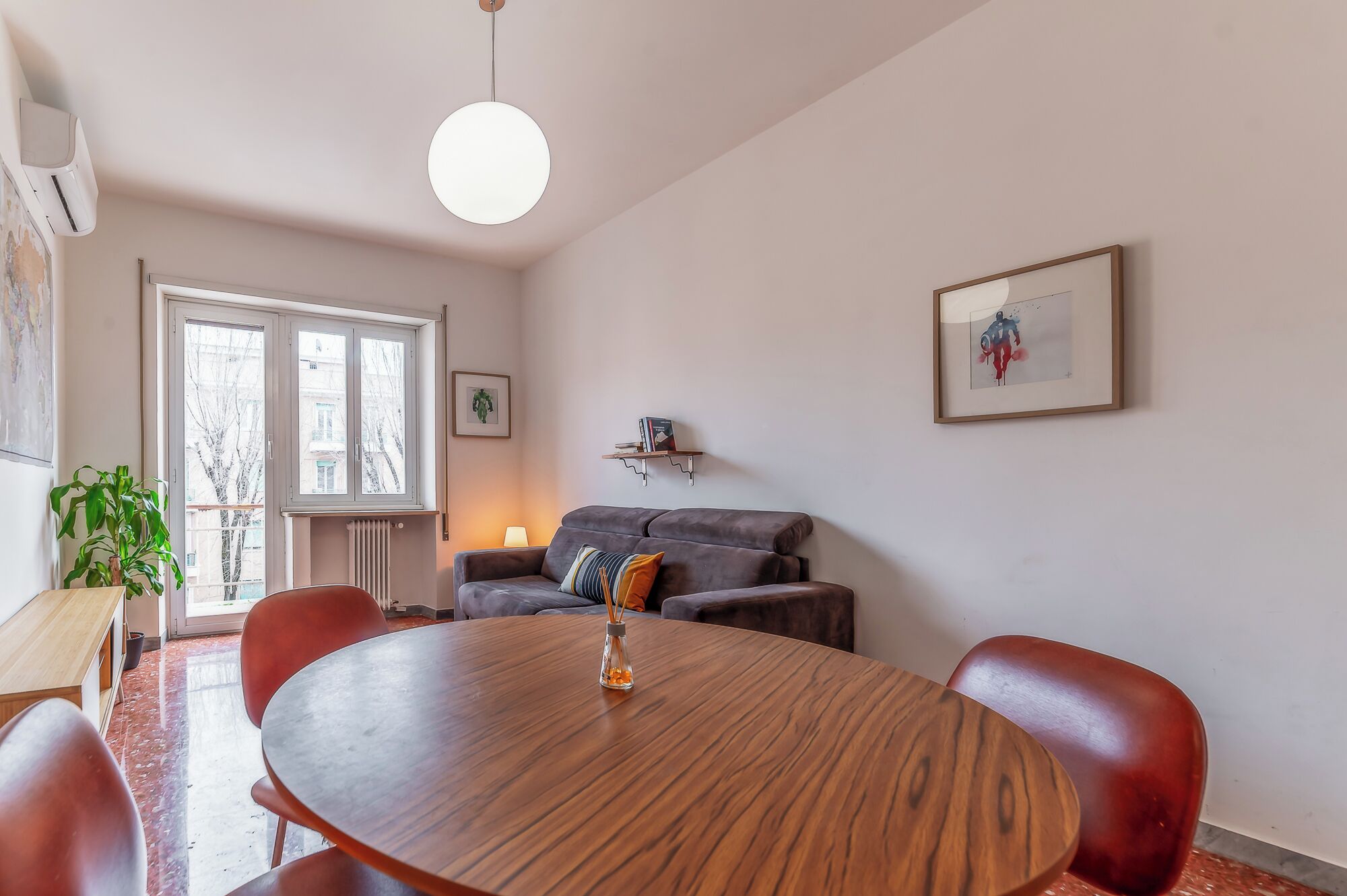 A dining area with a round wooden table, red chairs, a grey sofa, and a pendant light.