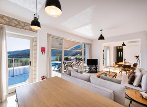 A modern living room with a large sofa, a dining table, and a TV. There is a balcony with a pool and a view of the hills. - Villa Cohili in Lefkada (Lefkada)