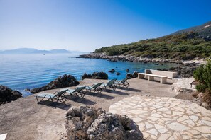 A coastal area with a stone patio, lounge chairs, and a rocky shoreline. - Villa Cohili in Lefkada (Lefkada)